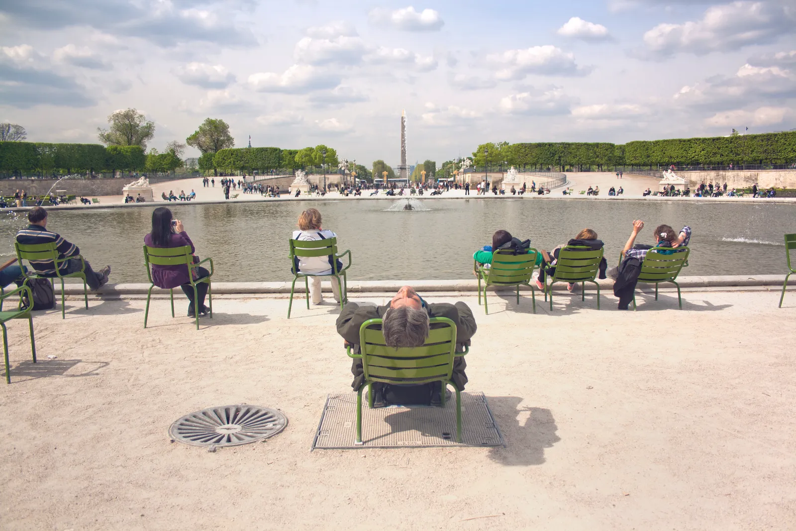 Visitors relaxing in green chairs beside the octagonal basin in the Tuileries Garden, with the Place de la Concorde obelisk visible in the distance