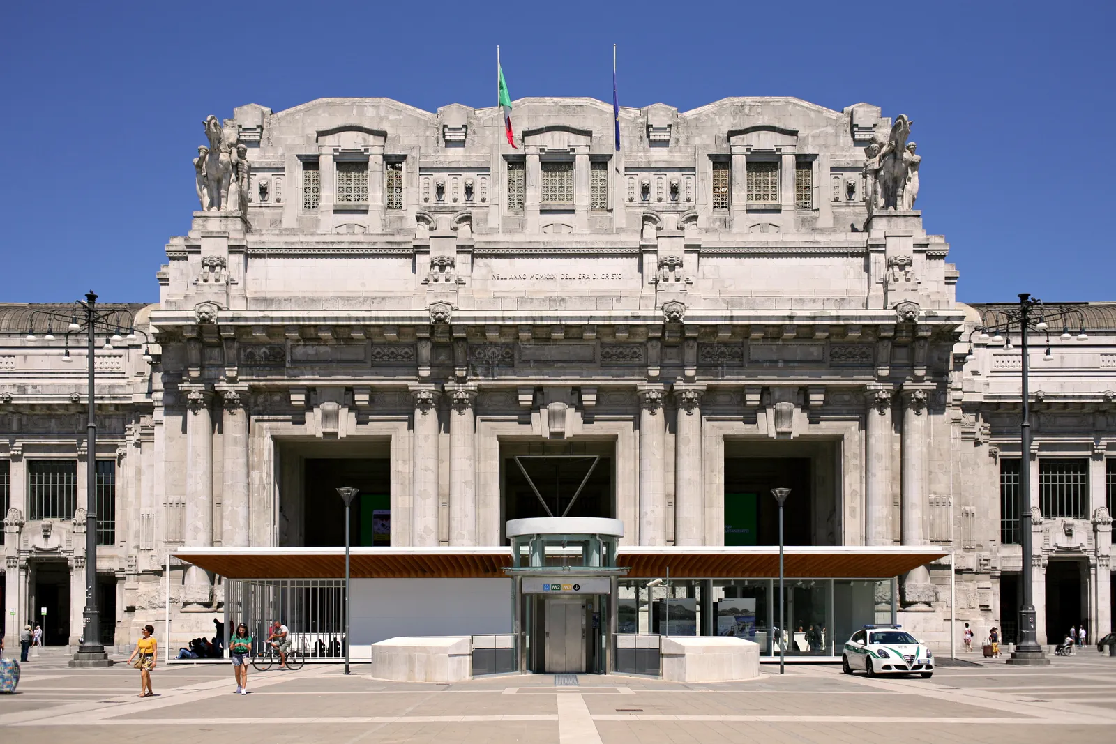 The monumental stone facade of Milano Centrale railway station, with equestrian statues and a wide plaza in front