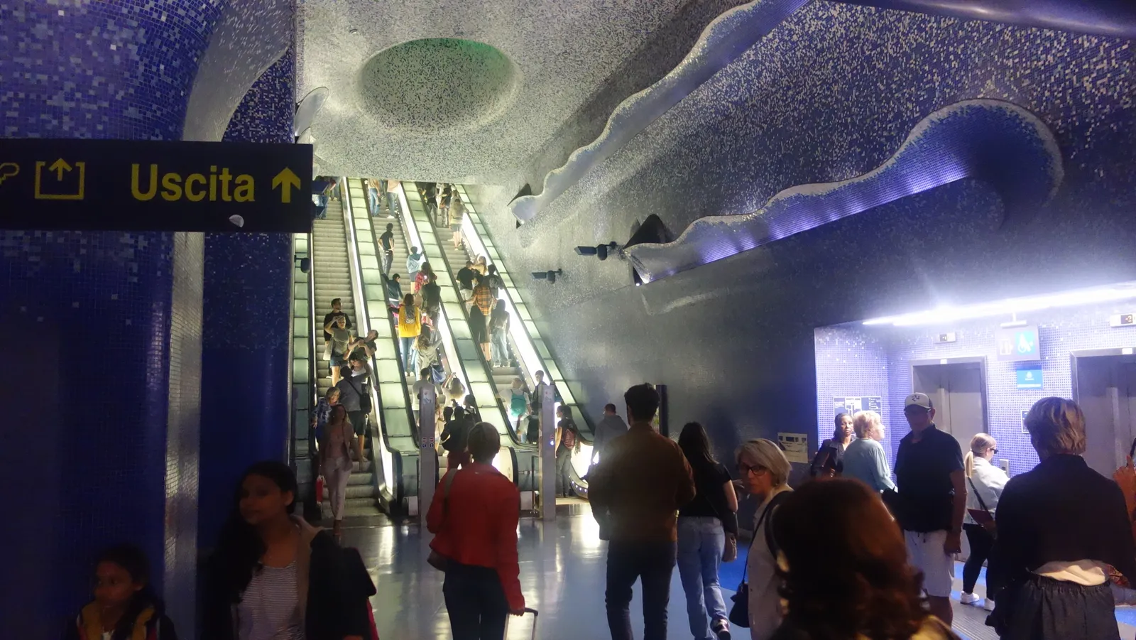 The blue mosaic mezzanine and escalators of Toledo metro station in Naples, crowded with passengers
