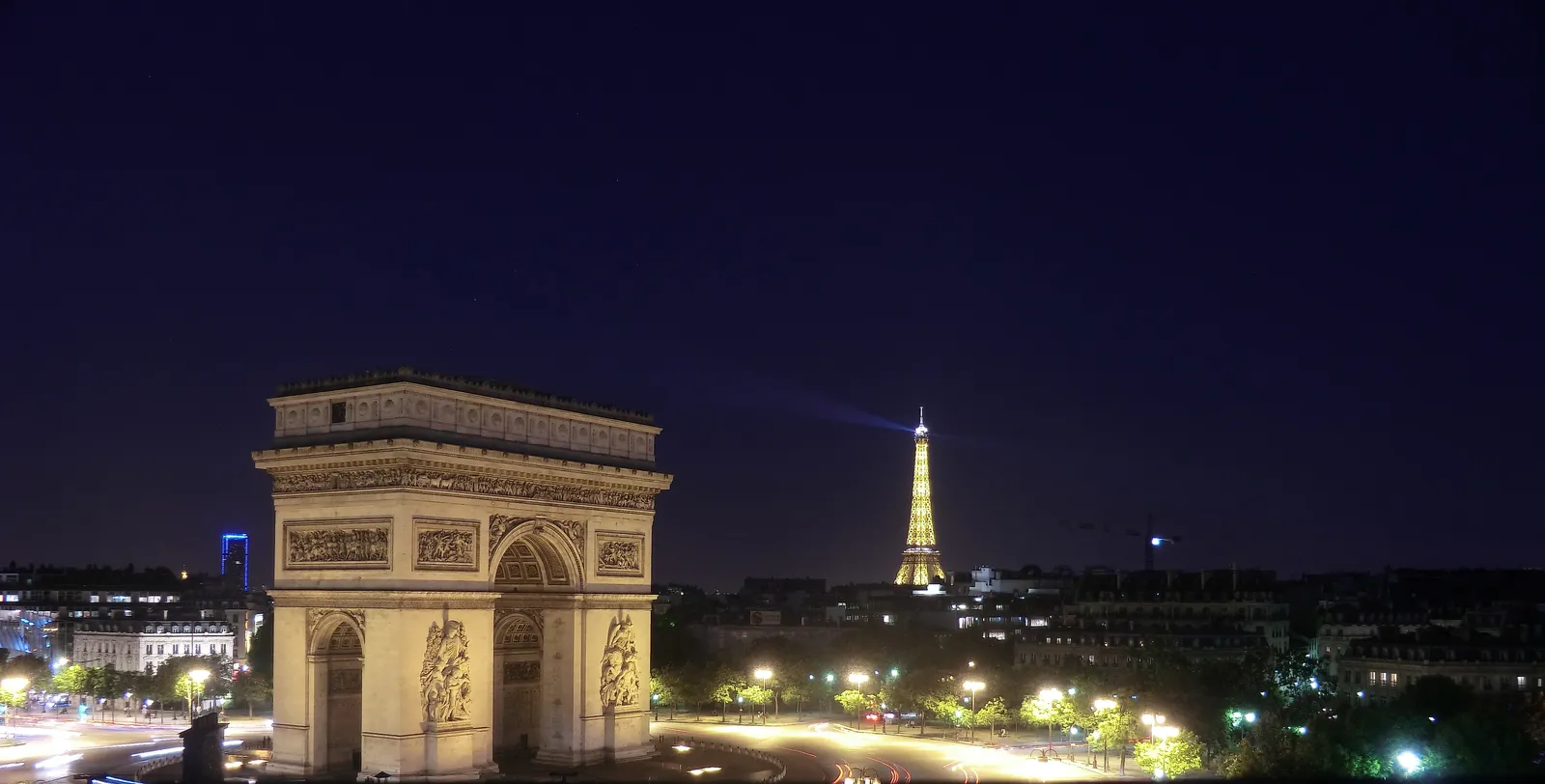 The Arc de Triomphe illuminated at night with the Eiffel Tower visible in the distance across the Paris skyline