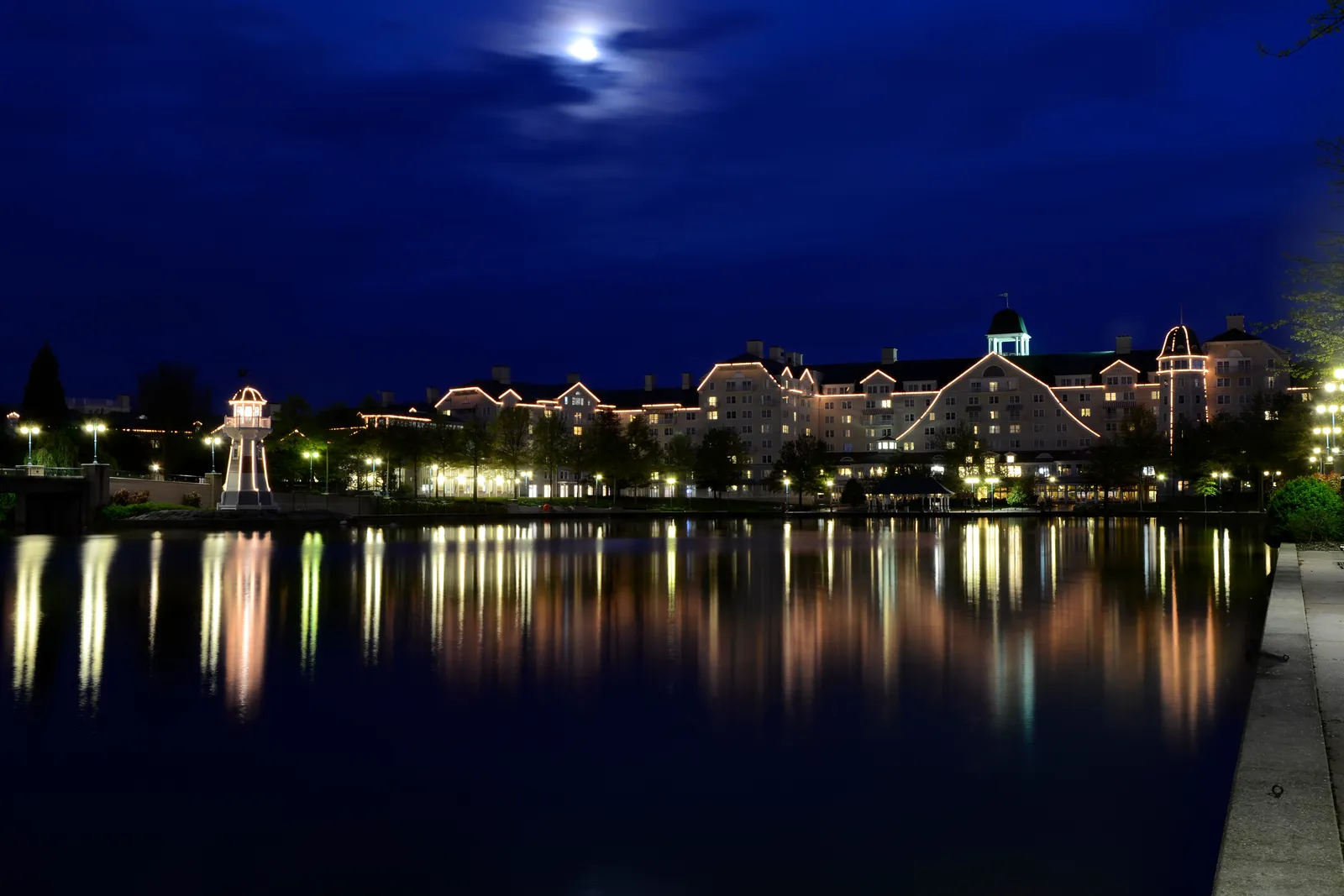 A lakeside resort hotel and lighthouse illuminated at night, reflected in the still water below