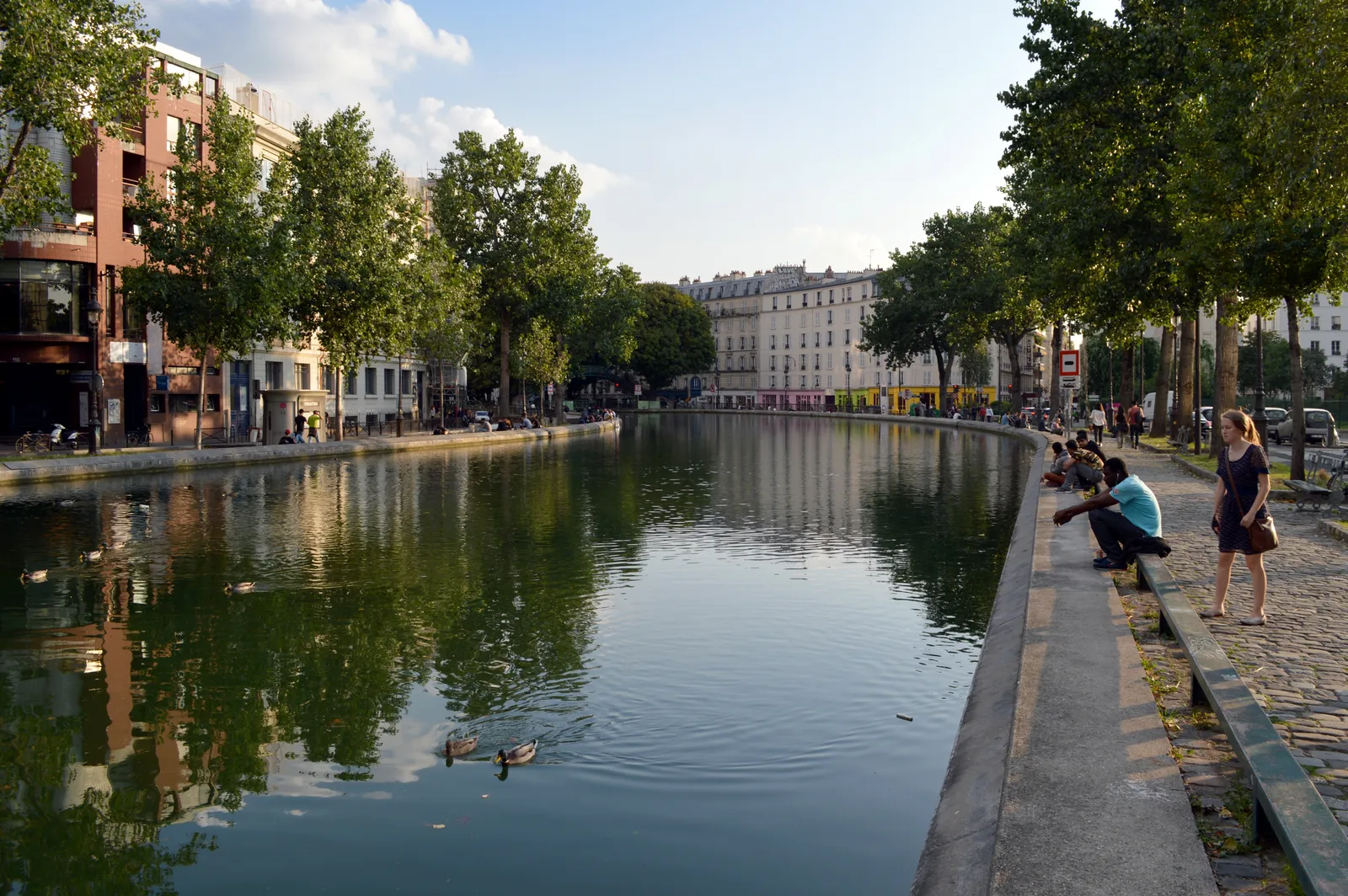People sitting along the tree-lined stone quay of Canal Saint-Martin with ducks on the still water