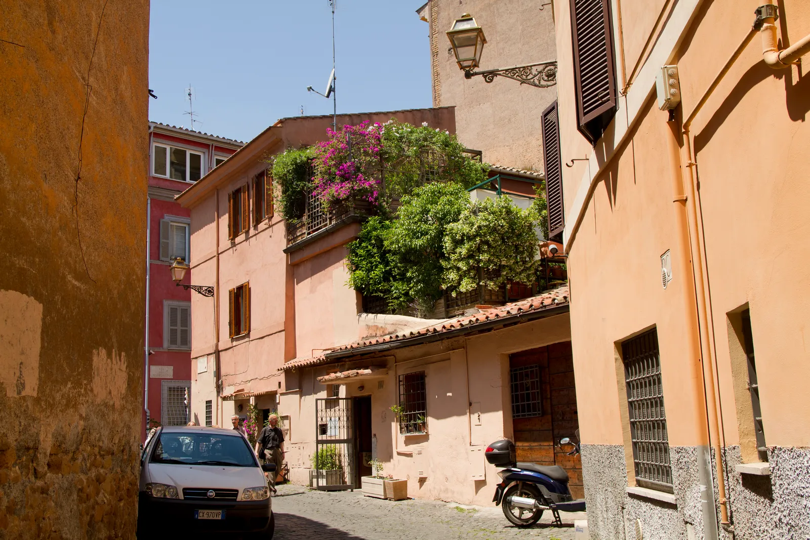A narrow Trastevere alley in Rome with ochre and pink buildings, bougainvillea flowers, and greenery spilling over a terrace
